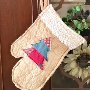 May include: Quilted oven mitt in a light tan color with a white, fluffy cuff. The mitt features a patchwork tree design in red, blue, and patterned fabrics. A loop is attached for hanging. The overall theme is rustic and handmade.