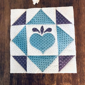 May include: A square quilt block featuring a central heart applique in teal with a small dark purple leaf motif above it. The design is surrounded by teal and dark purple triangles on a white background with a tiny blue floral pattern.