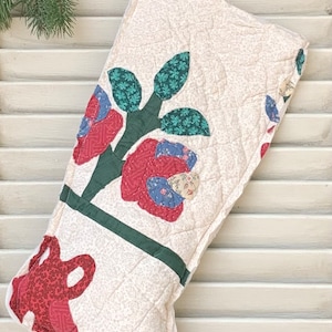 May include: A white quilted Christmas stocking with a red, green, and blue floral design. The stocking is hanging on a white wooden door.