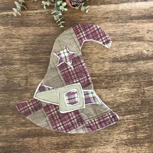 May include: A fabric witch hat ornament featuring a patchwork design of plaid and solid brown, tan, and maroon fabric squares. The hat includes a decorative band with a square buckle and a star-shaped patch, all finished with visible white stitching.
