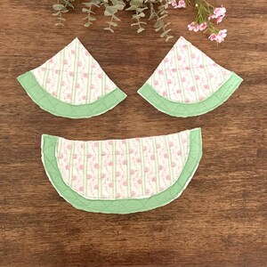 May include: Three quilted watermelon-shaped coasters. The coasters feature a white background with pink floral and green stripe patterns. Each coaster has a green border. The coasters are arranged on a wooden surface.
