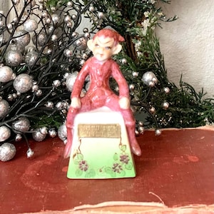 May include: A pink ceramic elf figurine sitting on a green and white base with the words "Don't Pity A-Doin' Look'y Like I'm Teasin'" written on it.