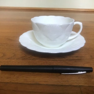 Könnte beinhalten: Ein weißes Tee-Set mit Untertasse und einem zarten Blumenmuster. Die Tasse hat eine abgerundete Form und einen Henkel, während die Untertasse einen gewellten Rand hat.