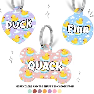 May include: Three pet tags with silver rings. One is heart-shaped with the word "DUCK" and a pattern of yellow rubber ducks on a purple background. Another is round with "Finn" and a blue background. The third is bone-shaped with "QUACK" and a pink background.