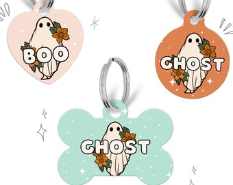 May include: Three different Halloween-themed dog tags featuring a ghost with flowers. The tags are in the shapes of a heart, circle, and bone. The heart tag is pink with the word "BOO" on it. The circle tag is orange with the word "GHOST" on it. The bone tag is light blue with the word "GHOST" on it.