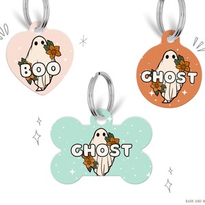 May include: Three different Halloween-themed dog tags featuring a ghost with flowers. The tags are in the shapes of a heart, circle, and bone. The heart tag is pink with the word "BOO" on it. The circle tag is orange with the word "GHOST" on it. The bone tag is light blue with the word "GHOST" on it.