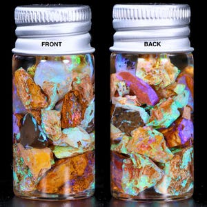 May include: Two small glass bottles filled with colourful, iridescent opal fragments. The bottles have silver lids and the text "FRONT" and "BACK" are visible. The opal pieces display orange, brown, and blue colours. The bottles are on a black background.