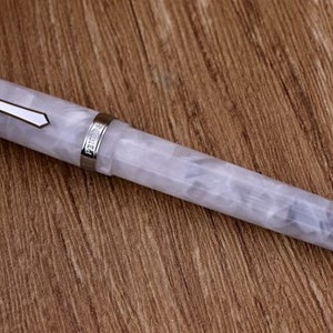 Può includere: Una penna stilografica bianca e grigia marmorizzata con dettagli argentati. La penna ha un design geometrico sul cappuccio e una fascia argentata vicino al pennino. Poggia su una superficie di legno.