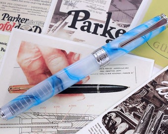 May include: A fountain pen with a translucent light blue and white marbled resin body, featuring a silver-toned metal clip and a wide engraved band. A slim black ballpoint pen lies underneath it, both resting on vintage paper advertisements.