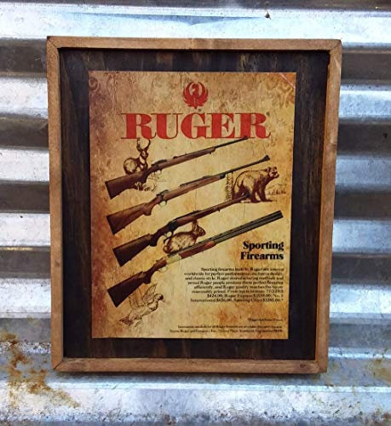 Puede incluir: Un anuncio vintage enmarcado para Ruger Sporting Firearms. El anuncio presenta un fondo de madera con una ilustraci&oacute;n en marr&oacute;n y blanco de varios rifles y animales. El texto "Ruger" est&aacute; en rojo y el texto "Sporting Firearms" est&aacute; en negro.