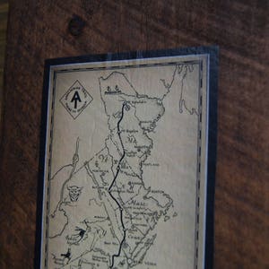 Op de afbeelding: Een ingelijste kaart van de Appalachian Trail, met een zwart-wit ontwerp en de tekst "Appalachian Trail" bovenaan. De kaart is op een bruine houten achtergrond.