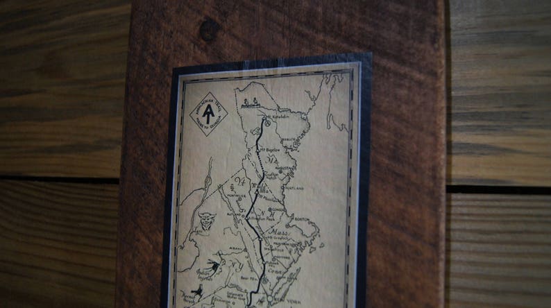 Op de afbeelding: Een ingelijste kaart van de Appalachian Trail, met een zwart-wit ontwerp en de tekst "Appalachian Trail" bovenaan. De kaart is op een bruine houten achtergrond.