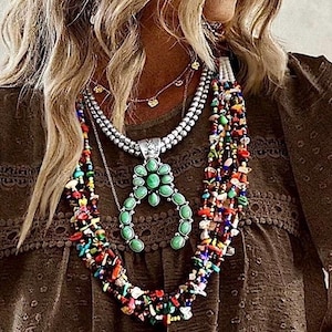 May include: A layered necklace set featuring a central silver-toned squash blossom pendant with green oval stones, a multi-strand necklace of small colorful chip beads, and a delicate silver-toned chain with small charms worn over a brown top.