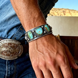 May include: A silver cuff bracelet featuring three oval turquoise stones with brown matrix, accented by twisted wire borders and small silver spheres. The bracelet is worn on a wrist alongside a large, ornate silver belt buckle with intricate engravings.