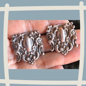 May include: A pair of silver earrings with an ornate, detailed design. The earrings are shaped like a heart with a raised oval in the center.