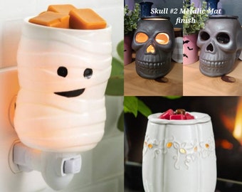 May include: Three Halloween-themed wax warmers. A white mummy-shaped warmer with a smiling face, a black skull-shaped warmer with a metallic finish, and a white pumpkin-shaped warmer with a floral design.