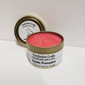May include: A gold candle tin with a red soy candle inside. The label reads "Purrfection Crafts Handmade with Love Apple Pumpkin 4.5 oz/127.5g Soy Candle."