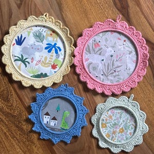 May include: Four round, decorative crochet frames in yellow, pink, blue, and light green. Each frame has a different patterned fabric insert with images of animals, houses, and flowers. The frames are placed on a wooden surface.