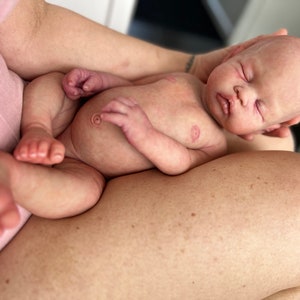 May include: A sleeping baby doll with closed eyes, rosy cheeks, and a soft, realistic appearance. The doll is lying on its back with its arms and legs slightly bent, and its head is tilted to the side. The doll is wearing no clothing.