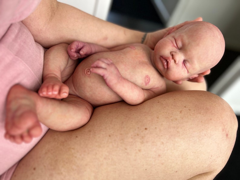May include: A sleeping baby doll with closed eyes, rosy cheeks, and a soft, realistic appearance. The doll is lying on its back with its arms and legs slightly bent, and its head is tilted to the side. The doll is wearing no clothing.