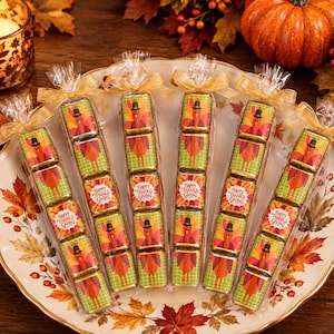 May include: Six clear cellophane bags containing square chocolate candies wrapped in foil. Each candy features a graphic of a turkey wearing a black hat or the text Happy Thanksgiving. The bags are tied with gold ribbons and arranged on a plate.