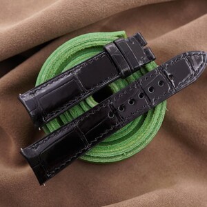 May include: A black watch strap with a glossy, textured finish and visible stitching along the edges. The strap features a buckle end with a loop and a pointed end with multiple circular holes for adjustment, resting on a coiled green band.