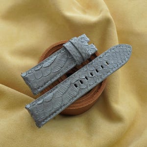 Puede incluir: Una correa de reloj gris de dos piezas con una superficie texturizada que imita escamas. La pieza más larga incluye una serie de orificios de ajuste rectangulares, mientras que la pieza más corta cuenta con una trabilla de cuero gris.