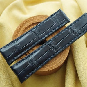 Puede incluir: Un par de correas de reloj de color azul marino oscuro con un patrón en relieve estilo caimán. Las correas están confeccionadas con costuras a juego en los bordes, presentando un acabado brillante y una forma rectangular y cónica.