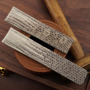 May include: Two watch straps made of gray leather with a textured pattern. The straps are lying on a wooden surface with a hammer and a piece of brown leather.