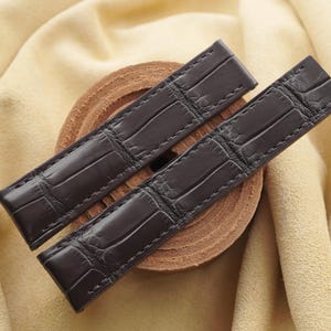 Könnte beinhalten: Ein dunkelbraunes Uhrenarmband mit einer strukturierten Prägung im Alligator-Stil. Das Armband besteht aus zwei separaten Teilen mit sichtbaren Nähten an den Kanten, einer glänzenden Oberfläche und einer rechteckigen Form mit zulaufenden Enden.