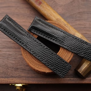 May include: Two black leather watch straps with a textured pattern. The straps are rolled up and placed on a wooden surface.