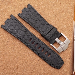 Puede incluir: Dos correas de reloj negras con un patrón texturizado similar a la piel de reptil y una hebilla plateada. Las correas están sobre una superficie marrón claro, junto a unas tijeras plateadas.