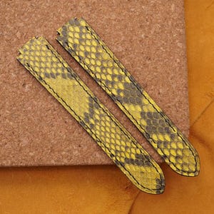 Peut inclure: Deux bracelets de montre avec un motif de peau de serpent jaune et gris. Les bracelets sont en cuir et ont des coutures noires. Ils sont présentés sur une surface marron avec un fond jaune.