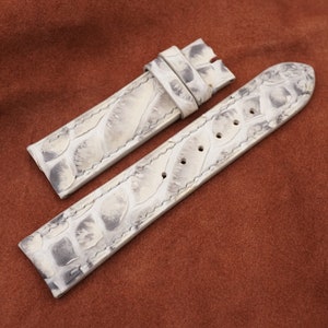 May include: Two white alligator watch straps with a textured pattern. The straps are laid out on a brown suede surface.
