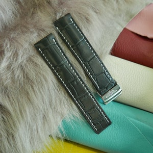 May include: Two black watch straps with a crocodile pattern and white stitching. The straps have a silver clasp with a logo. The straps are lying on a gray, furry surface.