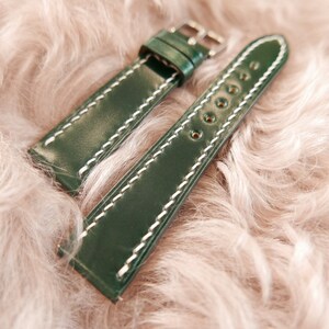 May include: A dark green leather watch strap with white stitching. The strap has a buckle and is shown on a fuzzy pink surface.