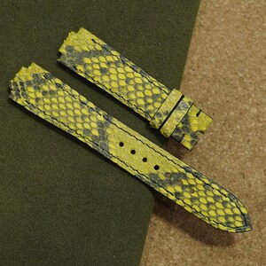 Puede incluir: Correa de reloj de dos piezas con un patrón de piel de serpiente amarilla y gris. La correa está hecha de un material texturizado y tiene un cierre de hebilla. La correa del reloj se muestra sobre una superficie verde y marrón.