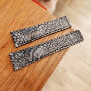 May include: Two gray leather watch straps with a textured pattern resembling scales. The straps are laid out on a wooden surface.