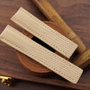 Puede incluir: Dos correas de reloj de cuero beige con un patrón texturizado. Las correas están sobre una superficie de madera.