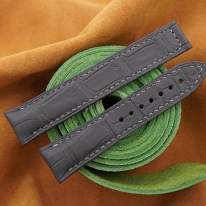 Peut inclure: Bracelets de montre gris avec un motif crocodile et des coutures blanches. Les bracelets sont présentés sur un ruban vert et un fond en daim beige. Les bracelets ont plusieurs trous pour le réglage.