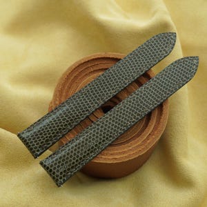 Könnte beinhalten: Zwei lange, spitz zulaufende Uhrenarmbänder in dunklem Olivgrün mit einem ausgeprägten, sich wiederholenden hexagonalen Schuppenmuster. Die Bänder liegen diagonal auf einem aufgerollten, hellbraunen Lederstreifen mit sichtbaren Kanten.