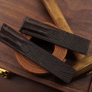 Könnte beinhalten: Zwei dunkelbraune Uhrenarmbänder mit einem reptilienartigen Muster liegen auf einer Holzoberfläche. Die Armbänder sind diagonal angeordnet, um ihr detailliertes Design zu zeigen. Der Hintergrund umfasst eine Holzkiste und Werkzeuge. Die Armbänder sind ca. 20 cm lang.