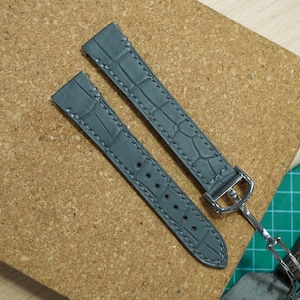Könnte beinhalten: Zwei graue Uhrenarmbänder mit Krokodilmuster, weißen Nähten und silberfarbenen Schnallen. Die Armbänder liegen auf einer Korkplatte, was auf einen Bastel- oder Reparaturrahmen hindeutet.
