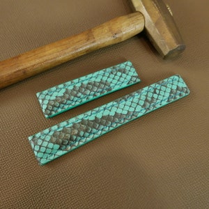 Puede incluir: Dos correas de reloj de cuero azul turquesa con un patrón de piel de serpiente. Las correas están dispuestas sobre una superficie de cuero marrón.