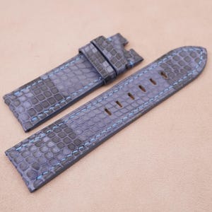 Puede incluir: Una correa de reloj de cuero azul con un patrón texturizado y costuras azules. La correa tiene una hebilla en un extremo y un conjunto de agujeros en el otro extremo.