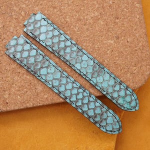 Peut inclure: Deux bracelets de montre avec un motif peau de serpent turquoise et gris. Les bracelets en cuir ont des coutures noires. Ils sont posés sur une surface marron avec un fond jaune.