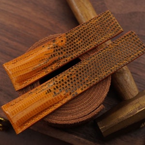 以下が含まれることがあります： 木製の表面に、ハンマーと茶色の革のロールと一緒に置かれた、テクスチャパターンが施されたオレンジ色の革製の腕時計ストラップ2本