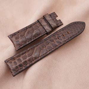 May include: A pair of brown watch straps made of textured leather, likely embossed to resemble reptile skin. The straps are in a neutral brown color and are designed to attach to a watch face. The background is a light tan color.