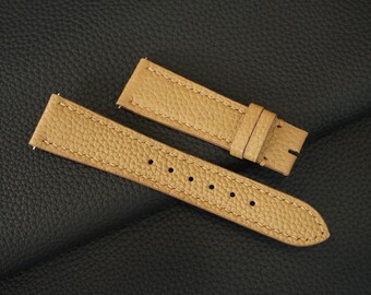 May include: Two tan leather watch straps with stitching. The straps are laid out on a black leather surface.