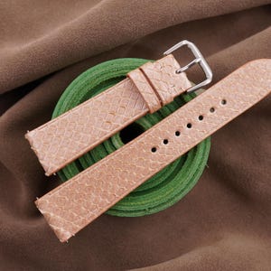 Könnte beinhalten: Hellbraune Armbanduhrenarmbänder mit strukturiertem Muster und silberner Schnalle. Die Armbänder werden auf einem grünen, kreisförmigen Objekt vor einem braunen Stoffhintergrund präsentiert. Die Armbänder sind ca. 20 cm lang.